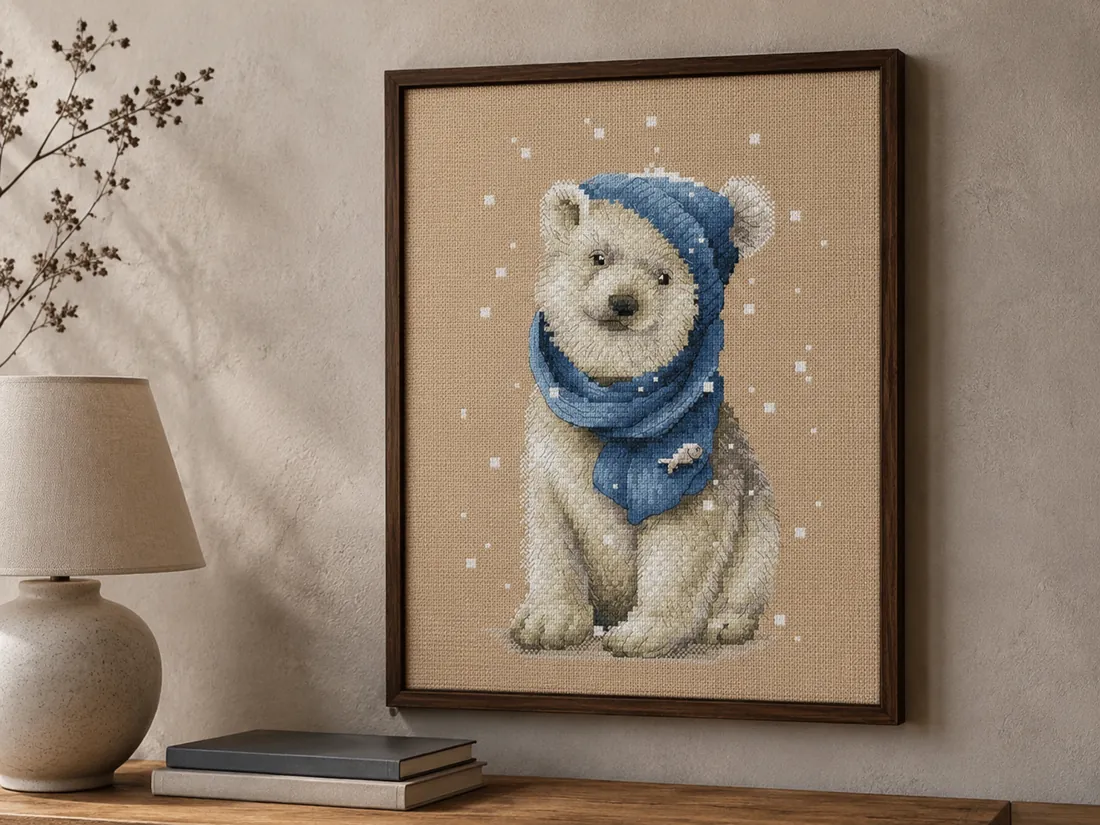 The finished White Bear design CC-K6-02 framed in dark brown wood on a gray-beige wall above an oak console, the polar bear cub in its denim-blue beanie and scarf on flaxen Aida